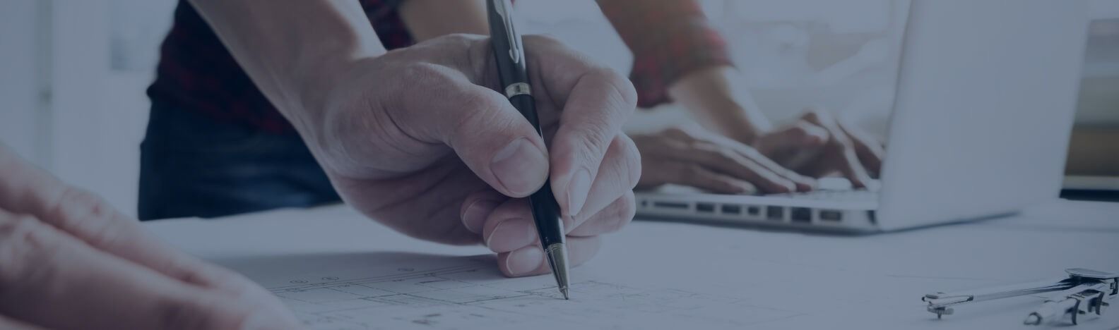 Man sieht eine Hand, die auf einen Bauplan zeichnet. Im Hintergrund sind zwei Hände, die auf einer Laptoptastatur tippen.